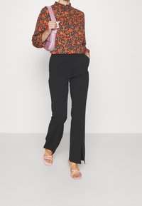 Blusa floreale multicolore con colletto arricciato, pantaloni neri sartoriali con spacco laterale e sandali rosa con strappo e accenti arancioni.
