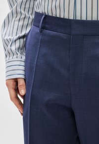 Παντελόνι σε ναυτικό μπλε με κομψή, λεία υφή, επίπεδη εμπρός όψη, πλαϊνές τσέπες και θηλιές για ζώνη, σε συνδυασμό με ριγέ πουκάμισο.