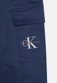 Marinblå cargobyxor med en ficklockslapp och vitt broderat "Calvin Klein Jeans"-logotyp med metalliska guldaccenter. Slät tygstruktur.