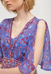 Fialové květinové šaty s krátkými volánkovými rukávy, detailním výstřihem a vzorovanou výšivkou v červených a modrých akcentů. Lehká, průhledná tkanina.