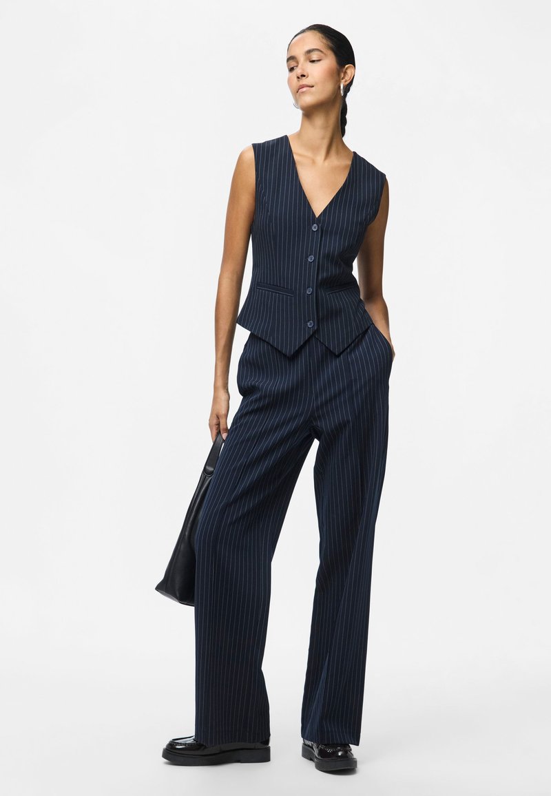 Chaleco ajustado a rayas marineras con un profundo escote en V y cinco botones, combinado con pantalones de pierna ancha a juego. Un tejido liso y estructurado.