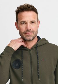 Sudadera con cremallera de color verde oliva con capucha acentuada en navy, presenta un logo circular y cordones, textura suave y corte casual.