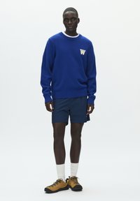 Pull en maille bleu avec col blanc et logo "W", associé à un short bleu marine et des chaussures de randonnée jaunes. Le mannequin se tient sur un fond blanc.