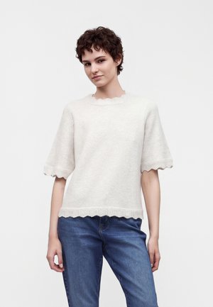 Femme aux cheveux courts et bouclés portant un pull blanc texturé à manches courtes avec des bords festonnés et un jean bleu sur fond uni.