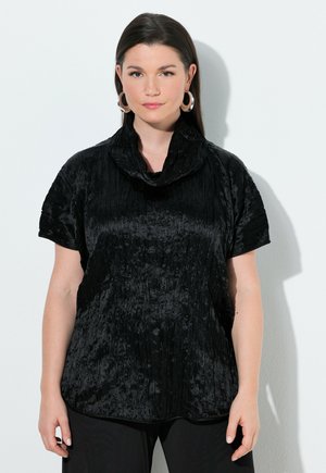 Kobieta o ciemnych prostych włosach, nosząca srebrne kolczyki koła i czarną, teksturowaną bluzkę z krótkim rękawem, stojąca na tle białej ściany.