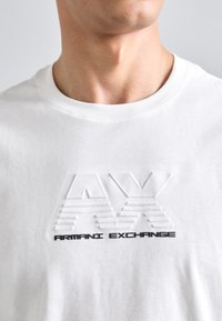 Valge puuvillane T-särk, millel on reljeefne "AX" logo ja must tekst "ARMANI EXCHANGE", ümar kaelus ja standartne lõige.