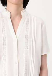 Žena má na sobě bílou halenku s krátkým rukávem, která má vertikální krajkové panely, jemné sklady, zapínání na knoflíky a volánkový stojáček.