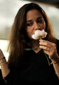 En kvinne holder en lys rosa blomst med grønne blader, iført en teksturert svart topp. Mykt lys skaper en mild atmosfære.