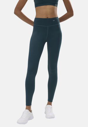 Mujer con leggins de cintura alta color verde azulado oscuro y zapatillas blancas, de pie con las manos detrás de la espalda sobre un fondo claro y liso.