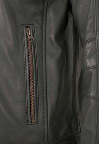 Dunkelgrüne Lederjacke mit einer seitlichen Reißverschlusstasche und bronzefarbenem Reißverschluss, glatter Textur und tailliertem Design mit genähten Akzenten.