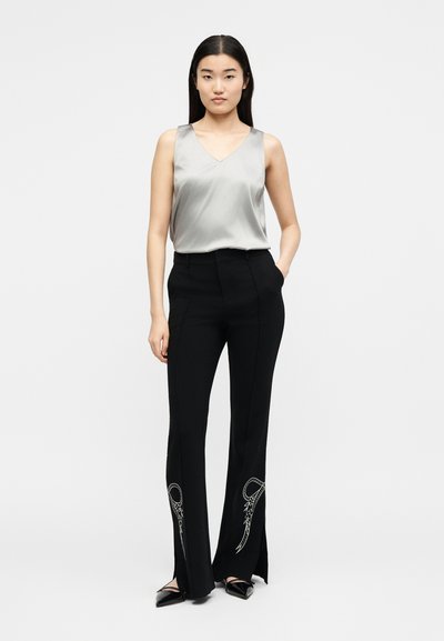 Μια γυναίκα στέκεται προς τα εμπρός φορώντας αμάνικη ασημένια σατέν μπλούζα και μαύρο παντελόνι με λευκά κεντημένα σχέδια κοντά στο τελείωμα.