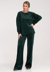 Zielony zestaw dresowy z weluru, składający się z luźnej koszuli z długim rękawem oraz spodni z szerokimi nogawkami, o gładkiej fakturze i delikatnym połysku.