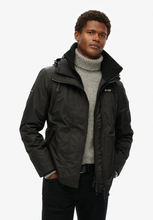 Chaqueta aislante negra con bolsillos con cremallera y un cuello alto, llevada sobre un suéter de cuello alto gris claro. Presenta un acabado de tela texturizada.