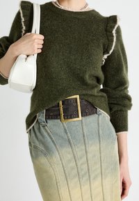 Femme portant un pull vert en tissu duveteux avec des épaules à volants, un collier de perles, une jupe en denim délavé avec des coutures verticales, et une large ceinture noire à boucle dorée.