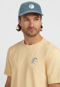 Lys gul t-shirt med korte ærmer og et cirkulært blåt og orange logo. Komplementeret af en blå kordfløjlskasket med et cirkulært logo.