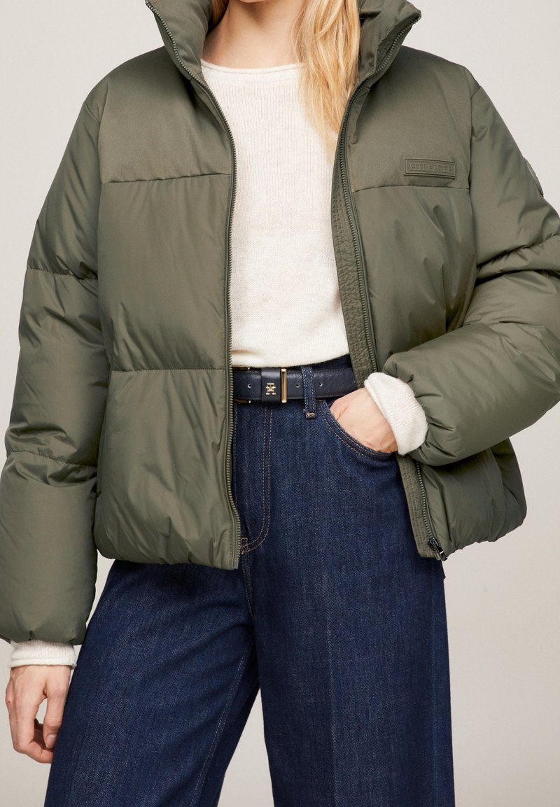 Personne portant une doudoune vert olive sur un pull crème, un jean bleu et une ceinture noire avec une boucle dorée.