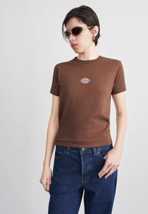 Persoană purtând un tricou maro Dickies, blugi albastru închis și ochelari de soare deschiși la culoare, pozând cu o mână în buzunar, pe un fundal simplu.