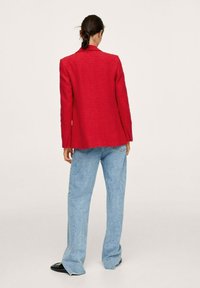 Blazer rouge texturé à coupe ajustée, associé à un pantalon en denim bleu clair taille haute et des chaussures plates noires. Vu de dos.