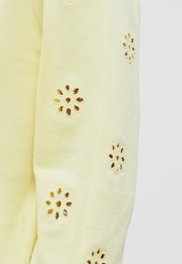 Lys gul sweatshirt med blomstermønster utskjæringer på ermene, med en myk tekstur og et avslappet design.