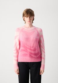 Jonge persoon met kort haar, gekleed in een roze gebreid trui met patroon en zwarte broek, staande tegen een effen witte achtergrond.
