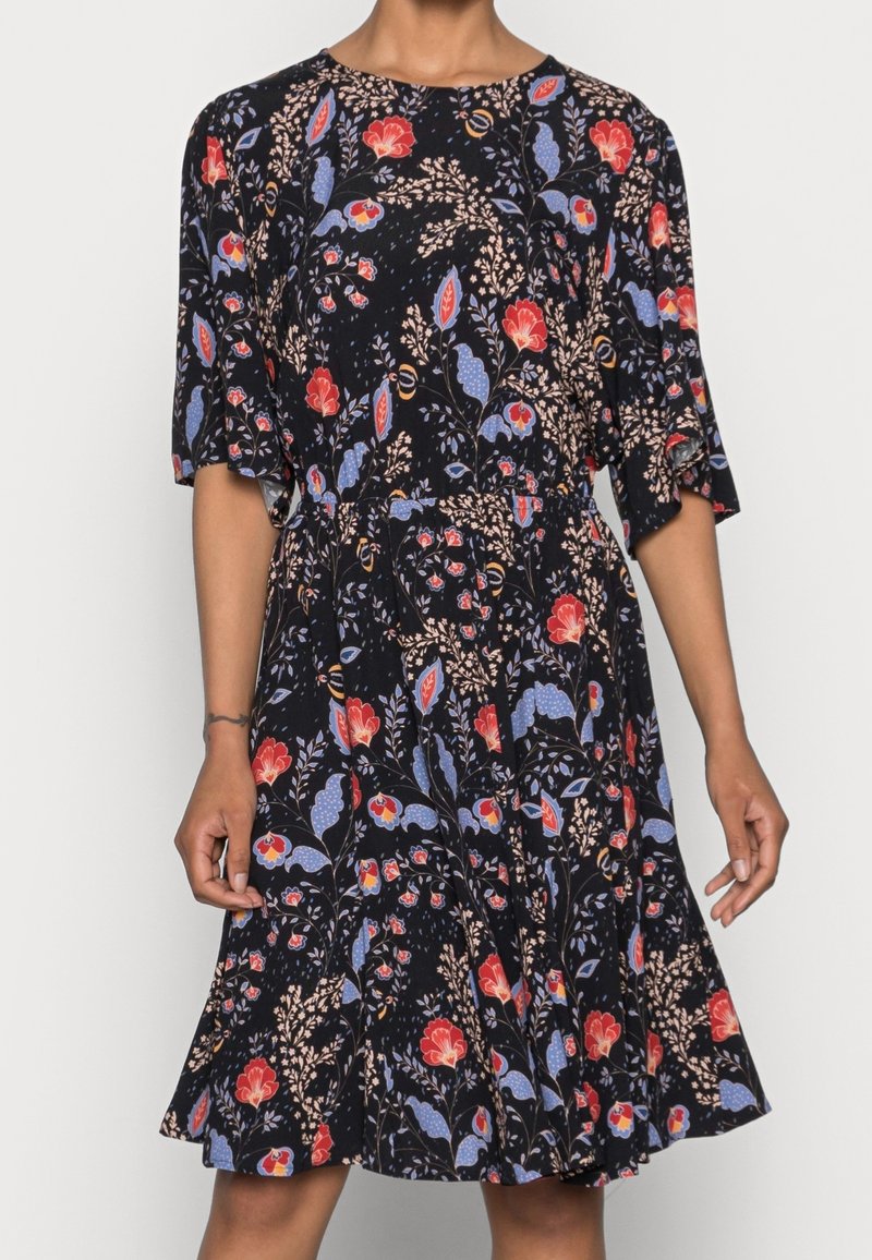 Femme portant une robe noire mi-longue avec des motifs floraux rouges, bleus et beiges et des manches jusqu'au coude.