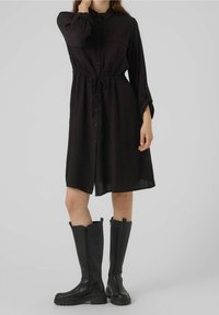 Abito camicia lungo nero con maniche lunghe, bottone frontale, vita con cordino e due tasche sul petto, abbinato a stivali alti neri con zip.