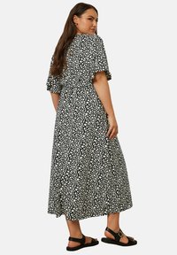 Svart maxiklänning med vitt abstrakt mönster, korta volangärmar, cinchad midja och svängig nederdel. Stylist med svarta sandaler.