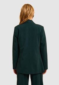 Blazer verde scuro con vestibilità aderente, revers a leva e una texture di tessuto liscia. Presenta una fessura singola sul retro per facilitare il movimento.
