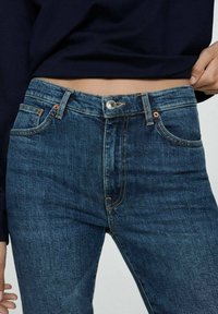 Jeans din denim albastru închis cu talie înaltă, dotate cu accesorii din cupru, design cu cinci buzunare și o ușoară decolorare în textura stofei.