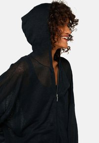 Haut à capuche zippé noir avec une texture transparente, des manches larges et une capuche doublée, présentant un design simple et fonctionnel.