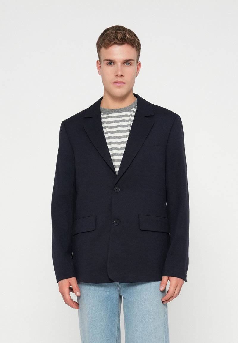 Blazer navy con rever a taglio, due tasche frontali e due bottoni. Indossato sopra una maglietta a righe grigie e bianche e jeans azzurri chiari.