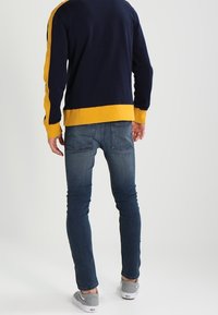 Marinblå och gul pullover-tröja med ribbad krage och muddar, kombinerad med mörk tvättade slim-fit jeans och grå slip-on skor.