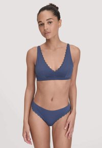 Vrouw met teruggetrokken haar, gekleed in een blauwe lingeriebra met kant en bijpassende onderbroek tegen een effen lichte achtergrond.