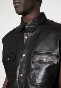 Svart läder ärmlös skjorta med spetshals, silverfärgade tryckknappar och två framfickor med klaffstängningar. Slät textur.