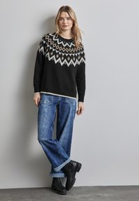 Svart stickad tröja med geometriskt mönster, tillsammans med blå vida jeans som är upprullade vid fållarna och svarta ankelskor.