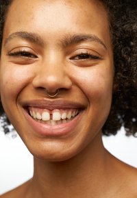 Nahaufnahme einer lächelnden Person mit lockigem schwarzen Haar, einem goldenen Septumring und heller Haut. Zu den Merkmalen gehören klare Zähne und ein natürlicher Teint.