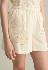 Pantalones cortos crema con una cintura elástica, que presentan bordados florales en los bolsillos. La tela es suave con un ligero brillo y un delicado detalle.