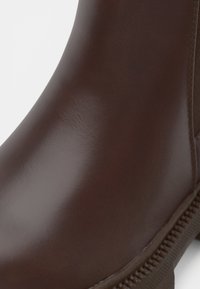 Close-up van een gladde donkerbruine leren laars met zichtbare stiksels langs de zool tegen een effen lichte achtergrond.