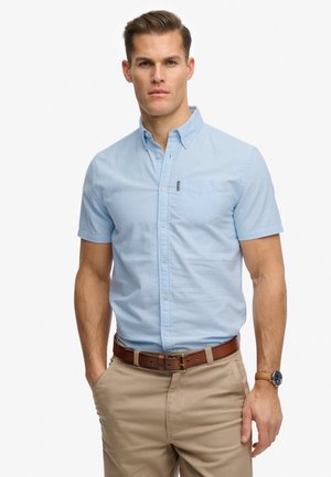 Mann trägt hellblaues Kurzarmhemd mit Knöpfen, beige Hose, braunen Gürtel und Armbanduhr, steht mit einer Hand in der Tasche vor weißem Hintergrund.