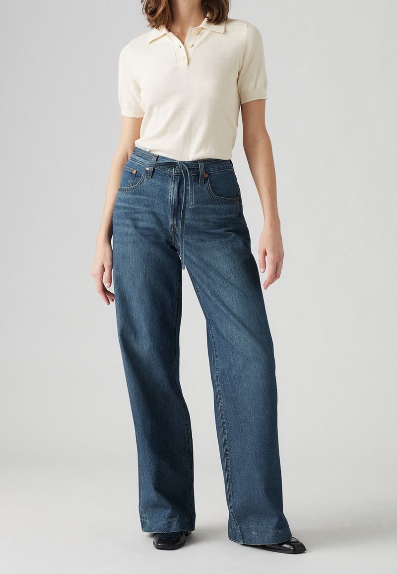 Femme portant un polo à manches courtes crème rentré dans un jean bleu taille haute à jambes larges avec une ceinture en tissu, se tenant contre un fond uni.