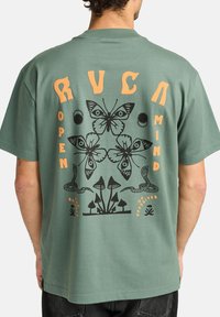Camiseta de algodón verde con un gran logo gráfico negro que presenta mariposas, setas y serpientes. El texto dice "MENTE ABIERTA" y "BALANCE".