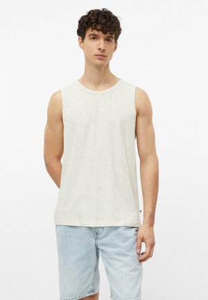 Junger Mann mit lockigem Haar, der ein hellbeiges ärmelloses Hemd und hellblaue Jeansshorts trägt, steht vor weißem Hintergrund.