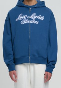 Sweat à capuche zippé bleu avec une poche kangourou, arborant le texte blanc "Low Lights Studios", des poignets côtelés et un tissu texturé.