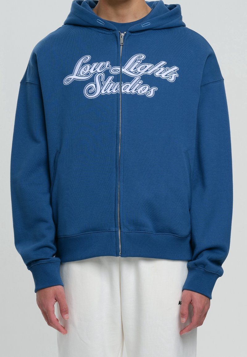 Sweat à capuche zippé bleu avec une poche kangourou, arborant le texte blanc "Low Lights Studios", des poignets côtelés et un tissu texturé.