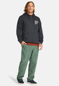 Man with wavy hair wearing a dark hoodie and green corduroy pants, standing confidently with a slight smile against a plain background.