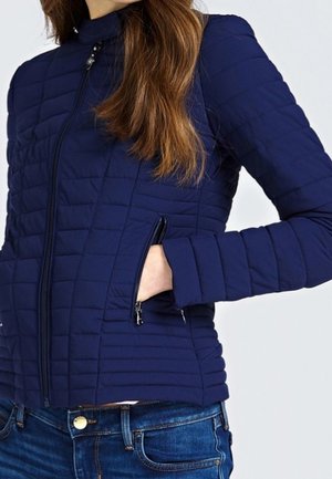 Woman wearing a navy blue quilted jacket with a front zipper and zippered pockets, paired with blue jeans, hand in pocket.