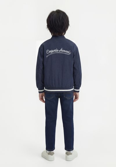 Παιδί που στέκεται με την πλάτη, φοράει ναυτικό μπλε μπουφάν Emporio Armani, μπλε τζιν, λευκά αθλητικά παπούτσια, σε απλό λευκό φόντο.