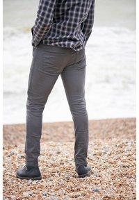 Gray slim-fit pants with a smooth texture, paired with black ankle boots. The individual stands on a pebbled beach.