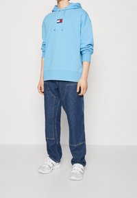 Ljusblå hoodie med logotypapplikation, tillsammans med mörkblå jeans med unik sömnad, och vita sneakers.