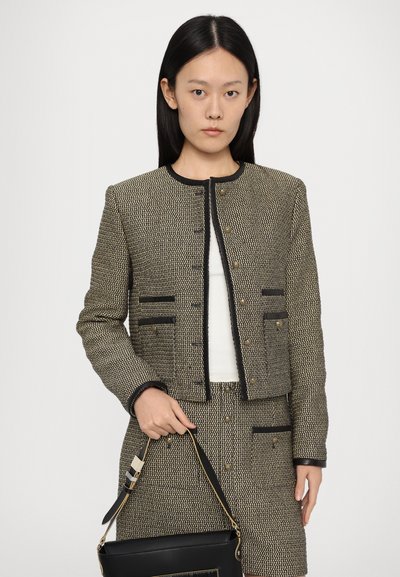 Tweed jacket in black and beige, cropped design with button closure, patch pockets, and black trim. Model holds a black handbag.